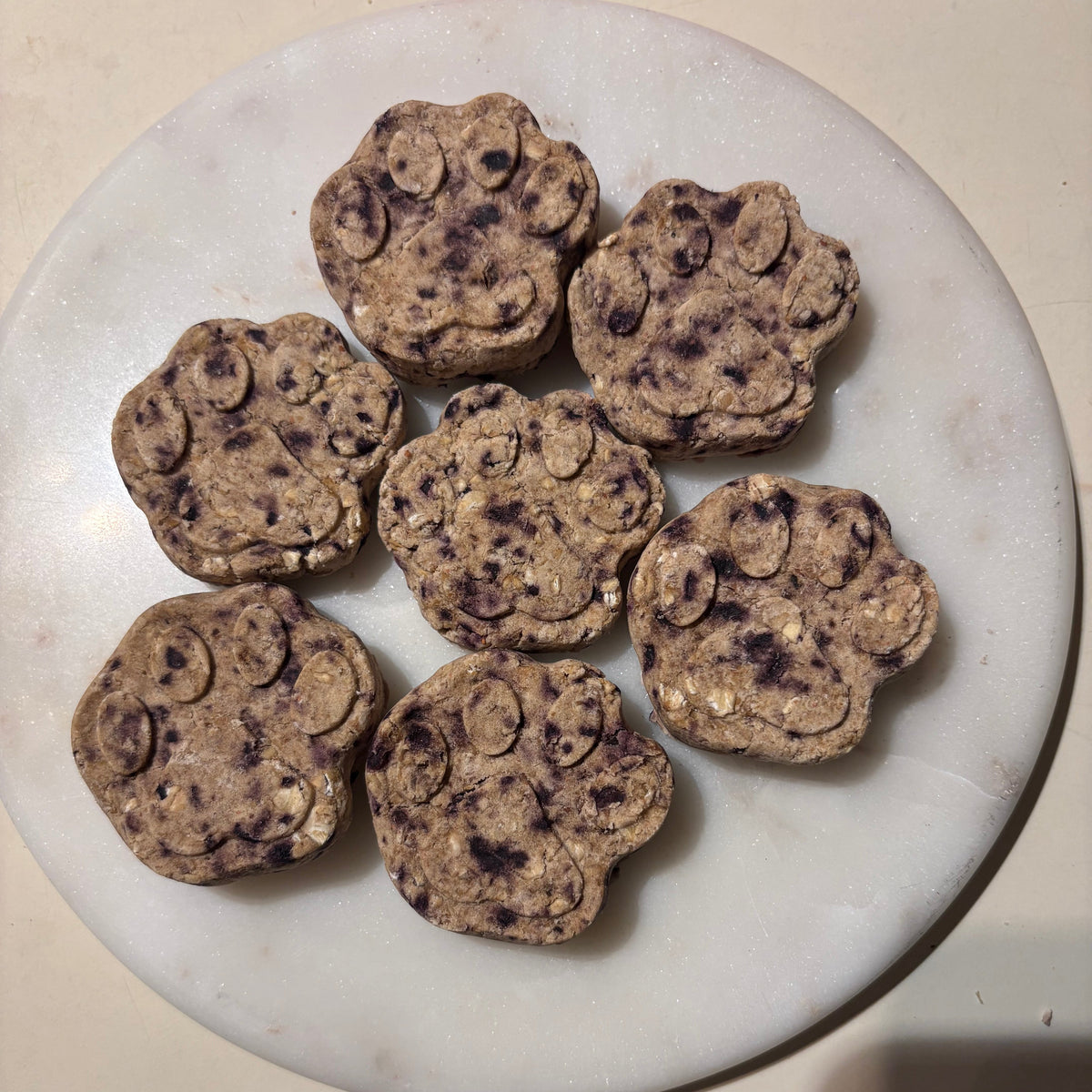 Six paw-shaped dog treats with embedded dark purple dried blueberry pieces, arranged on a round white marble serving board.