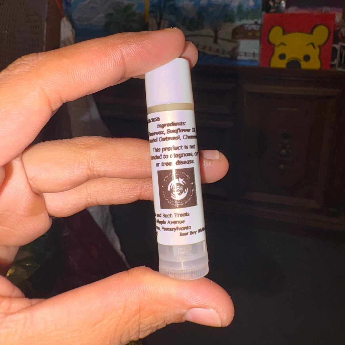A hand holding a small white lip balm tube with a printed label, photographed in a dim indoor setting with shelves and decor in the background.