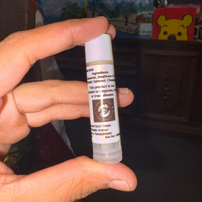 A hand holding a small white lip balm tube with a printed label, photographed in a dim indoor setting with shelves and decor in the background.